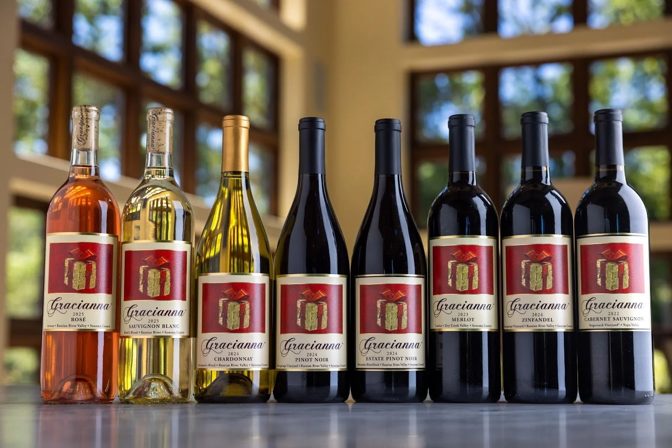 Seven bottles of wine from Gracianna winery, including rosé, sauvignon blanc, chardonnay, pinot noir, estate pinot noir, zinfandel, and cabernet sauvignon, lined up on a table with a wooden interior and large windows in the background.