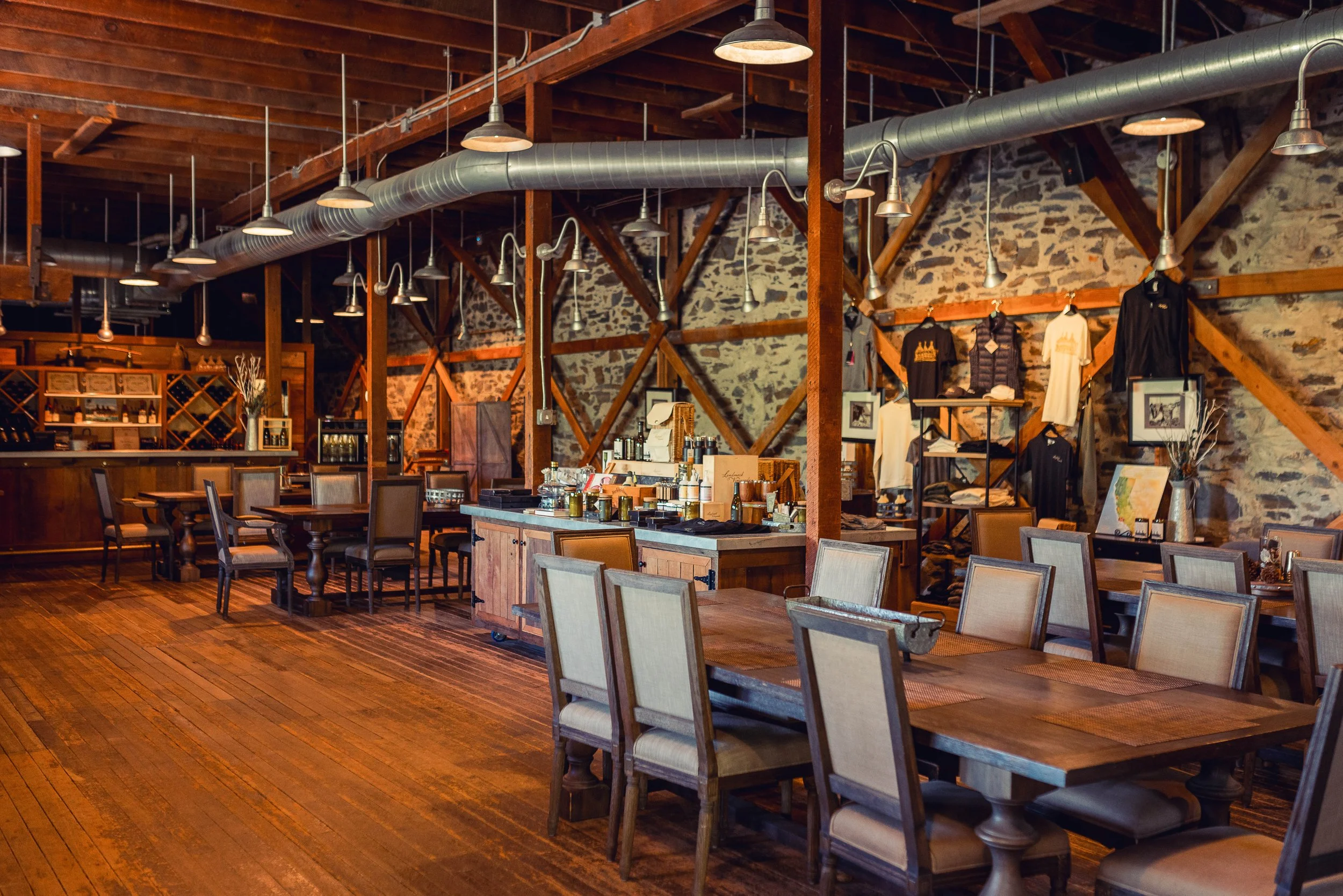 Interior of a rustic restaurant or cafe with wooden floors, exposed brick walls, and wooden beams on the ceiling. There are tables and chairs arranged throughout the space, with some shelves displaying bottles and artwork. Overhead, there are large i