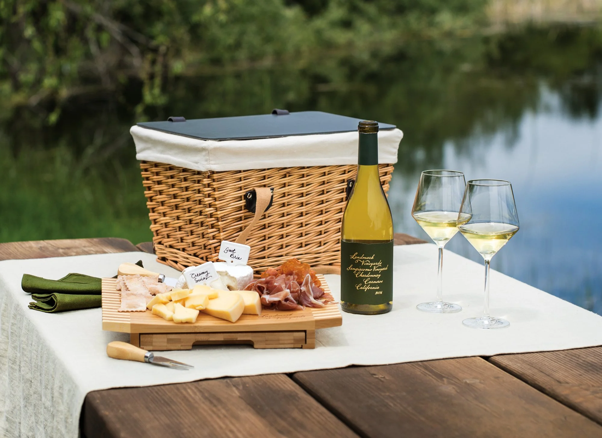 A picnic setup near a body of water with a wicker picnic basket, two glasses of white wine, a bottle of white wine, and a wooden cheese platter with assorted cheeses and cured meats on a white tablecloth.