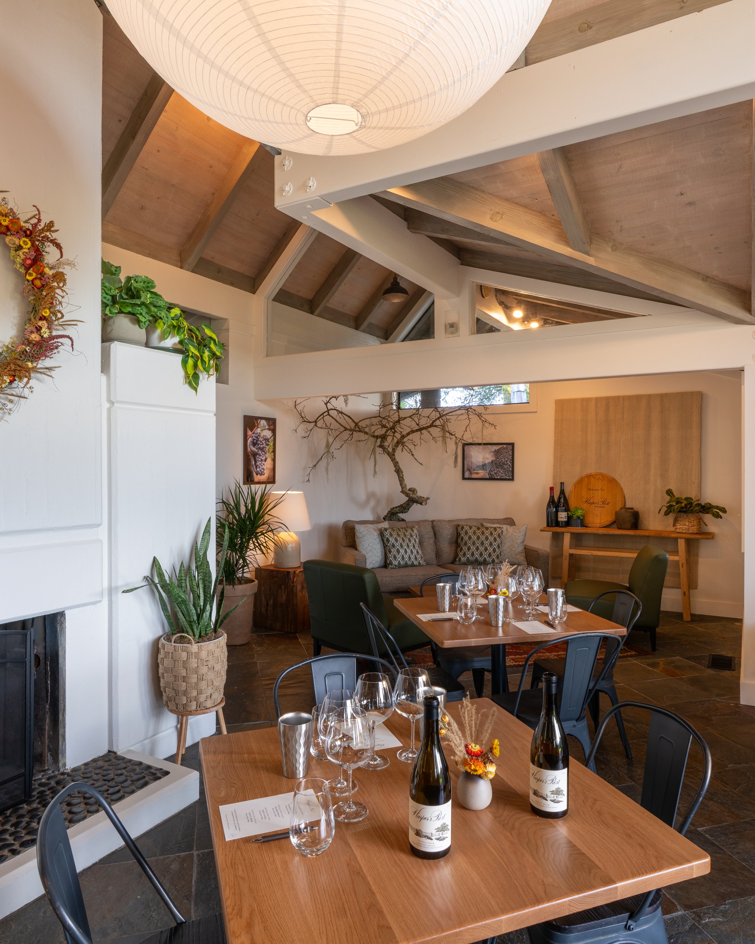 A cozy dining area in a restaurant featuring a wooden table set with wine bottles, glasses, and a small floral arrangement. There are black chairs around the table, a tan sofa with patterned pillows along the wall, and decorative plants including a l