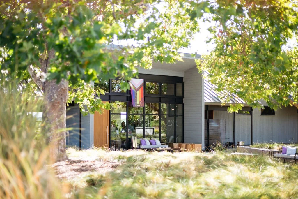 Modern house with large glass windows surrounded by trees and greenery, with outdoor seating area.