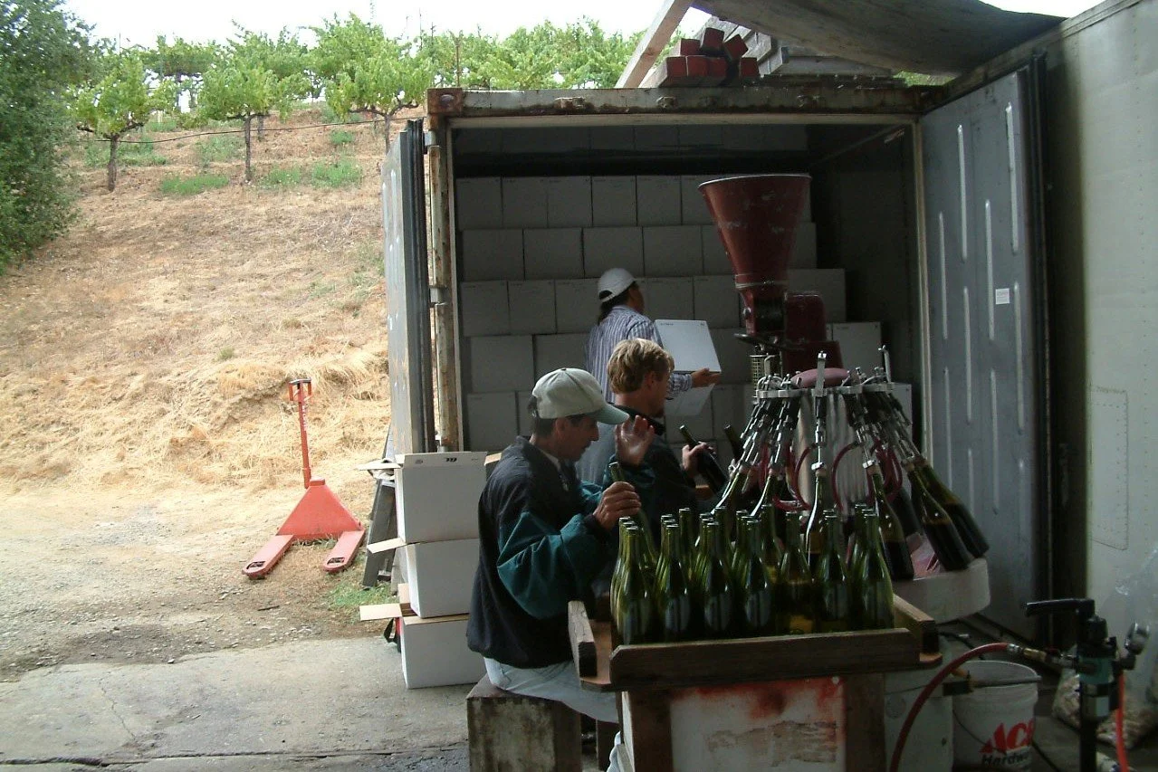 People working with bottles in a mobile wine bottling unit, with a vineyard on a hill in the background.