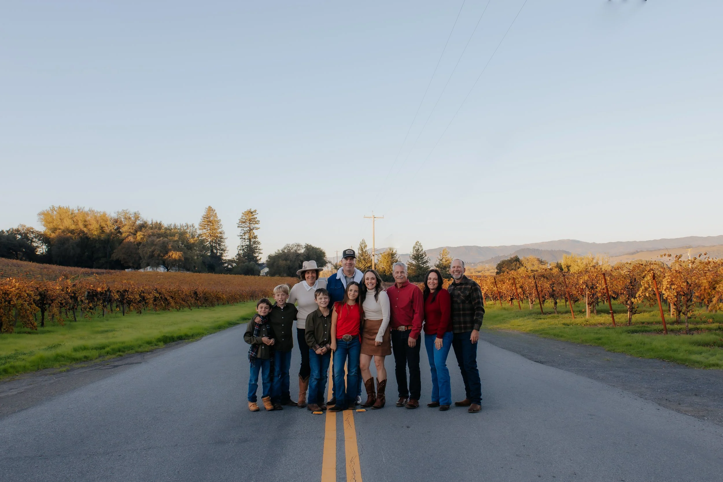 The Bacigalupi Family standing on a road in front of a vineyard with autumn foliage, hills in the background, under a clear sky.