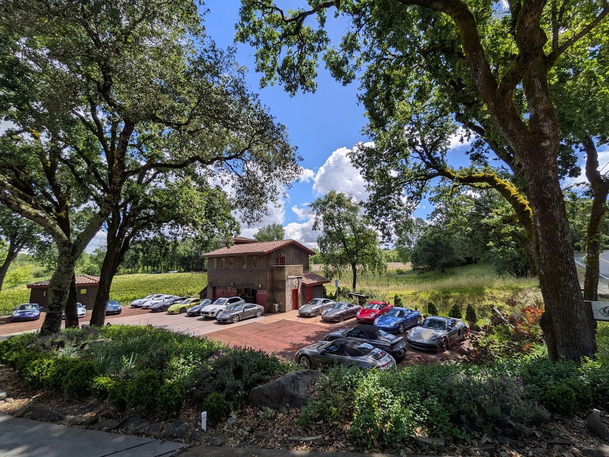 A parking lot with several cars parked in front of the Gracianna Winery building surrounded by tall trees and greenery under a partly cloudy sky.