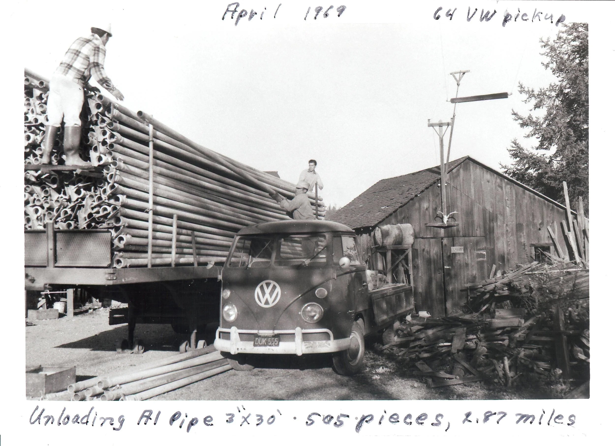 Black and white photo of a flatbed truck with a Volkswagen logo, carrying metal pipes, with two men working on the pipes. Handwritten notes indicate the date April 1, 1969, and the activity of unloading pipes, 3 inches by 30 feet, with five pieces to
