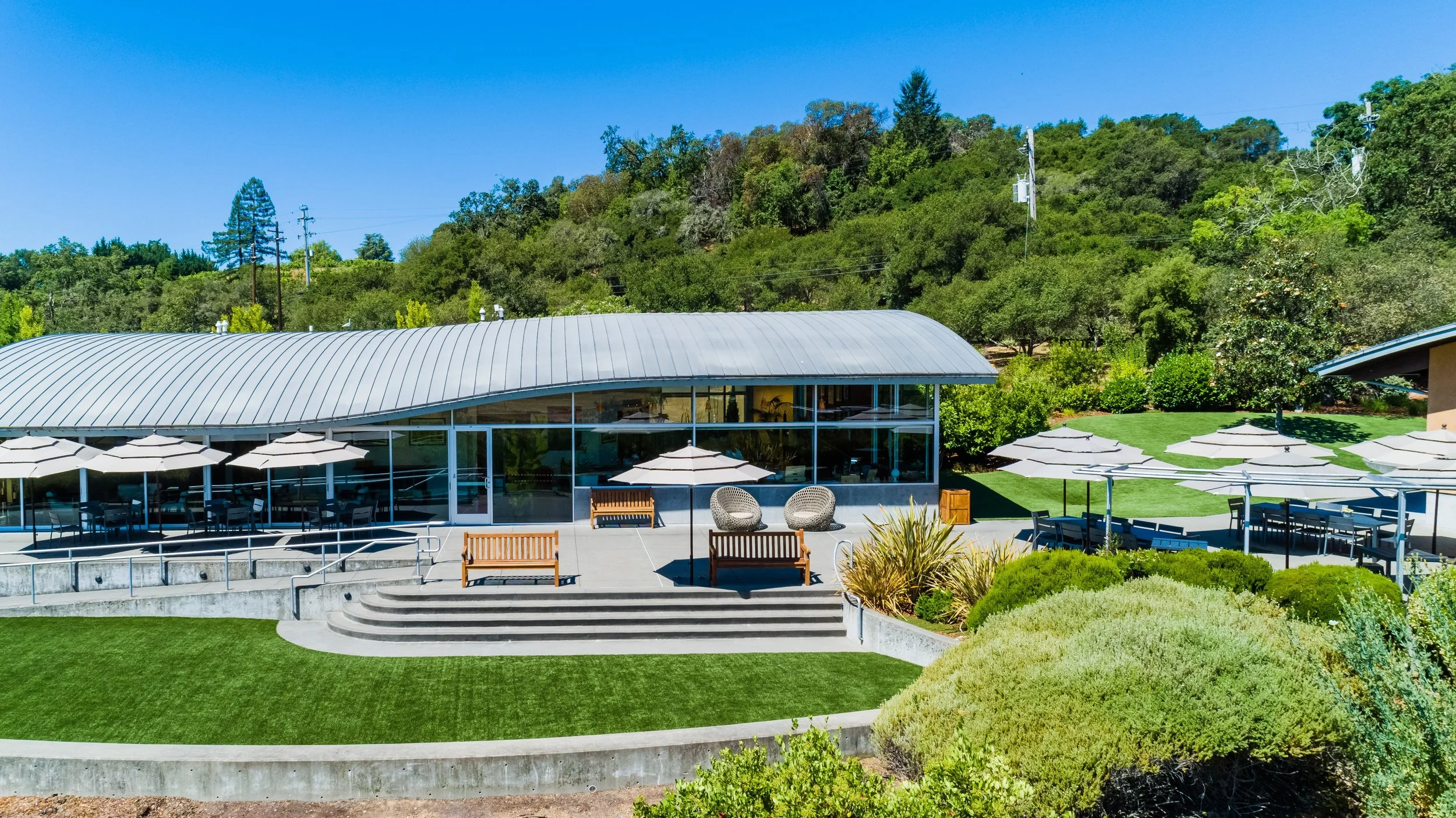 A building with a curved metal roof and glass walls, surrounded by a patio with umbrellas and outdoor seating, set against a hillside with trees and clear blue sky.