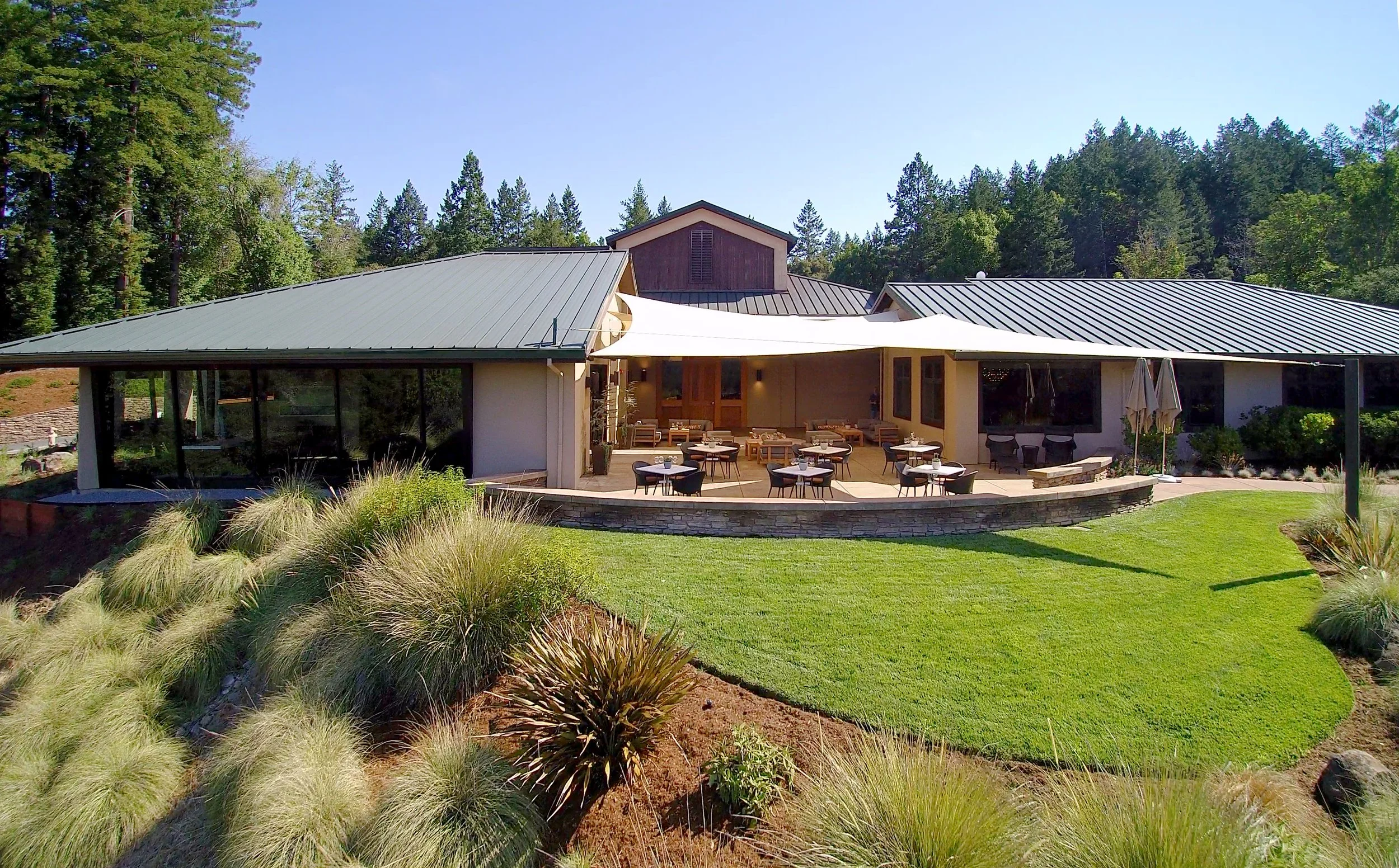A modern house with a metal roof and an outdoor patio area, surrounded by green grass, plants, and trees under a clear blue sky.