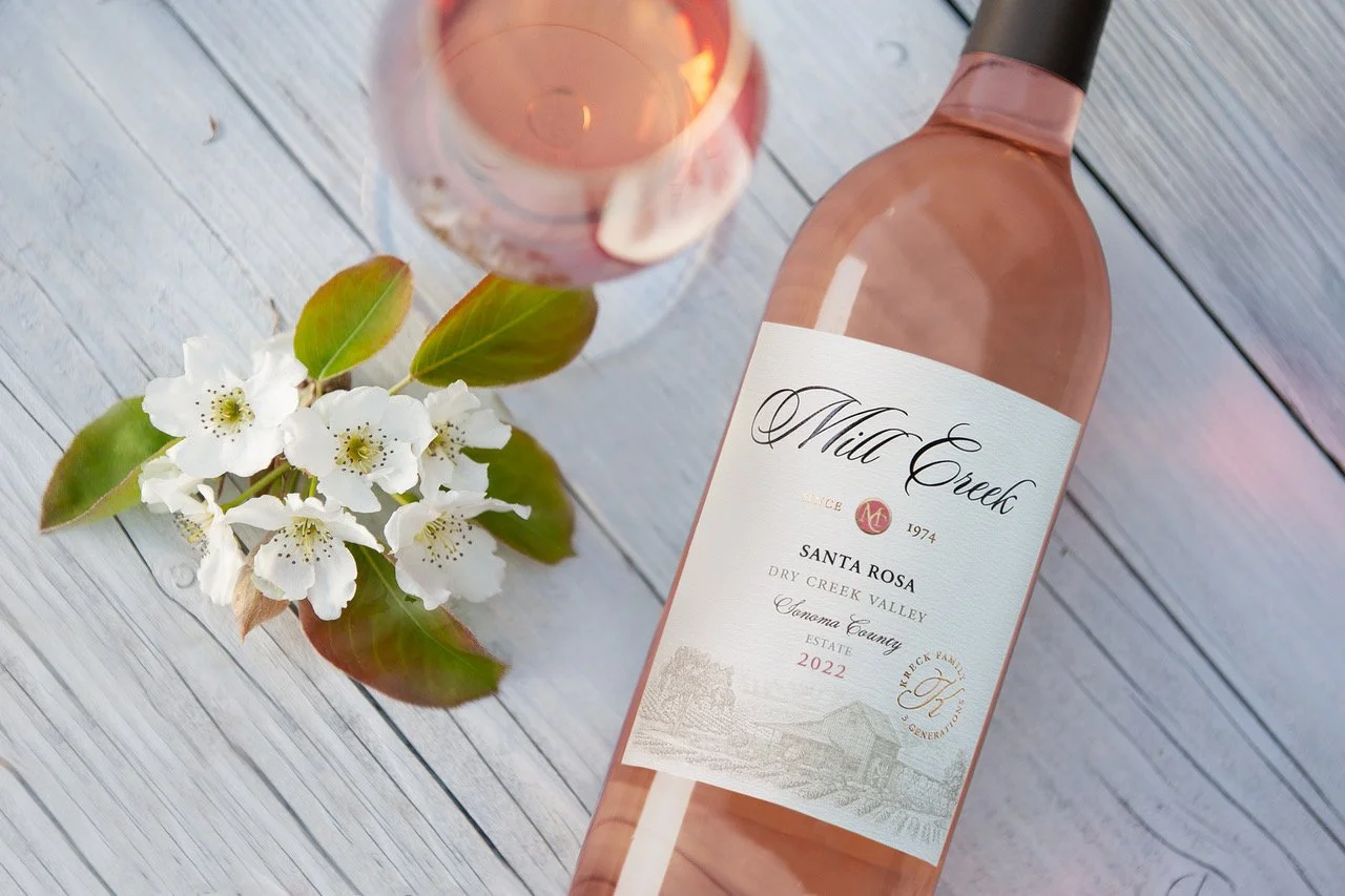 A bottle of rosé wine labeled 'Mia Creek', a glass of rosé wine, white blossoms with green leaves, and a white wooden surface.
