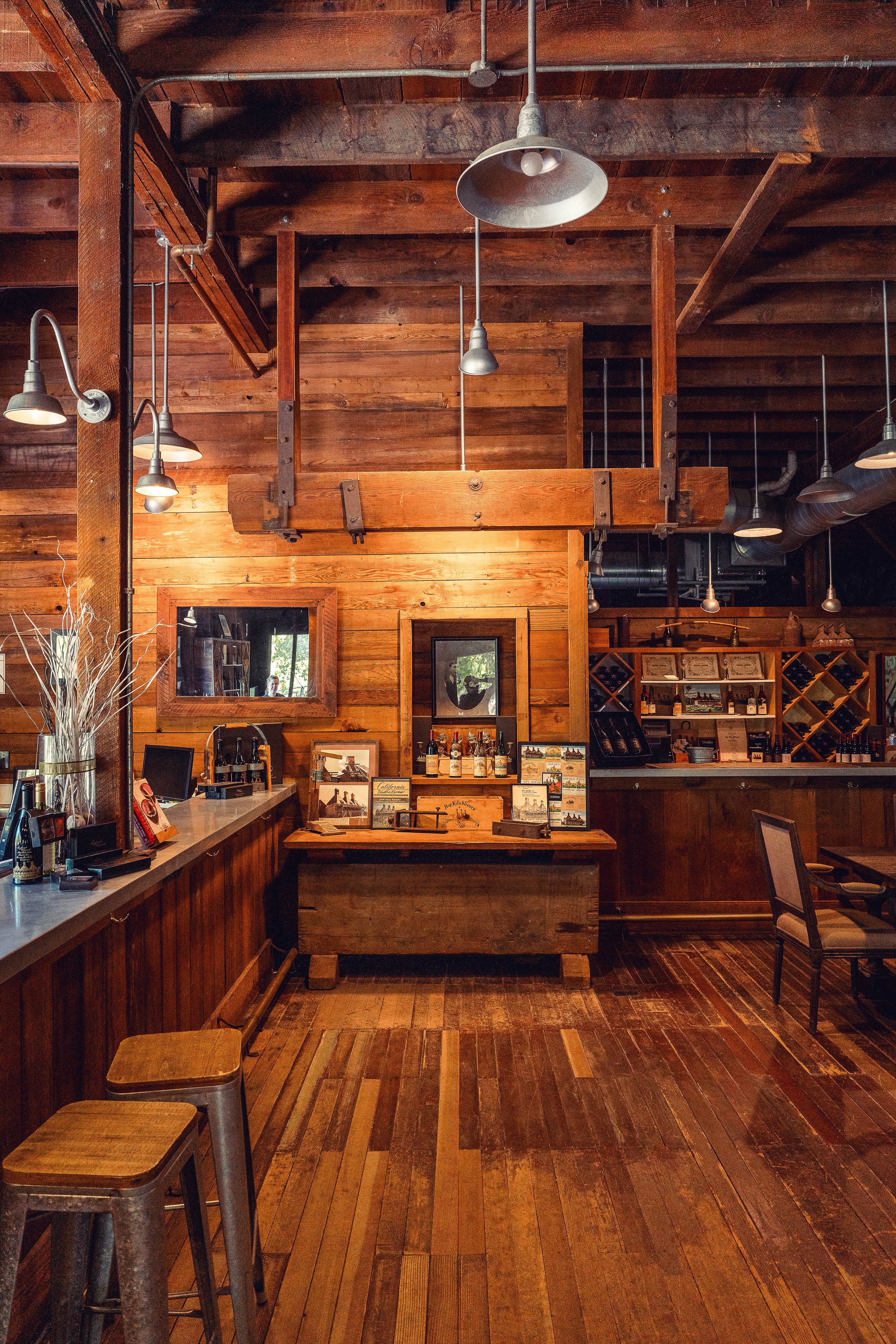 Interior of a rustic wooden winery or tasting room with a bar area, wine bottles, and decorative framed pictures, illuminated by hanging industrial-style lamps.