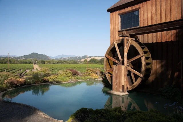 A wooden water mill beside a pond with scenic fields and hills in the background.