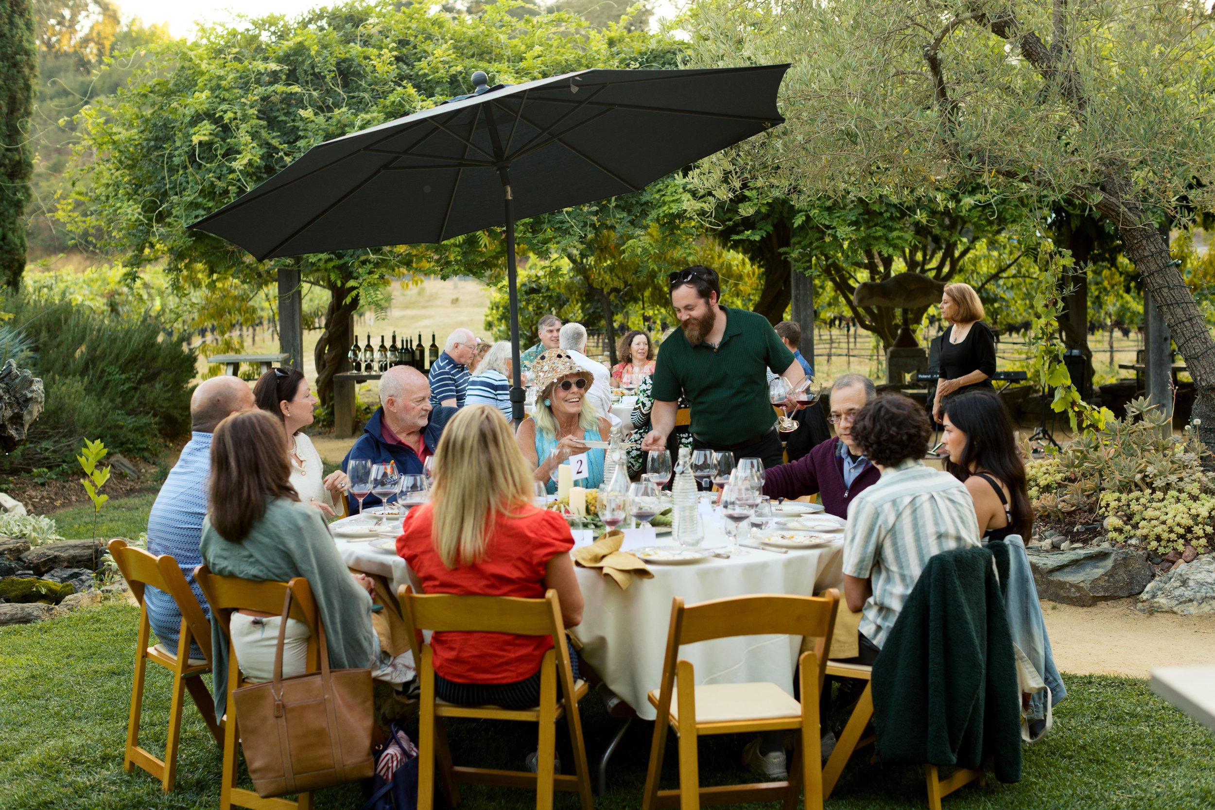 People gathered around outdoor dining tables in a garden setting, enjoying a meal with wine glasses, under a large black umbrella, surrounded by trees and greenery.