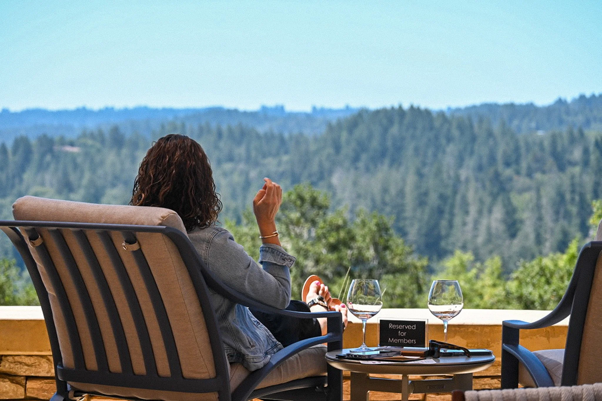 A woman with curly brown hair relaxing in a lounge chair on a terrace overlooking a forested landscape with hills.