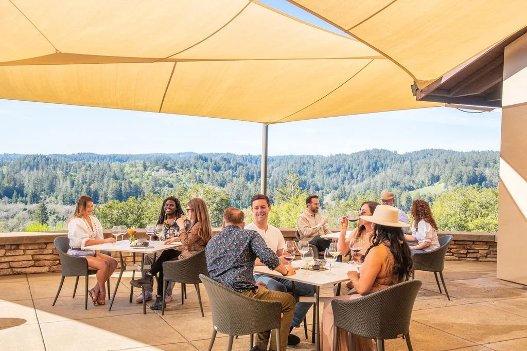 People tasting wine on a scenic patio at Gary Farrell Winery with a scenic view of green hills and trees in the background under a yellow canopy.