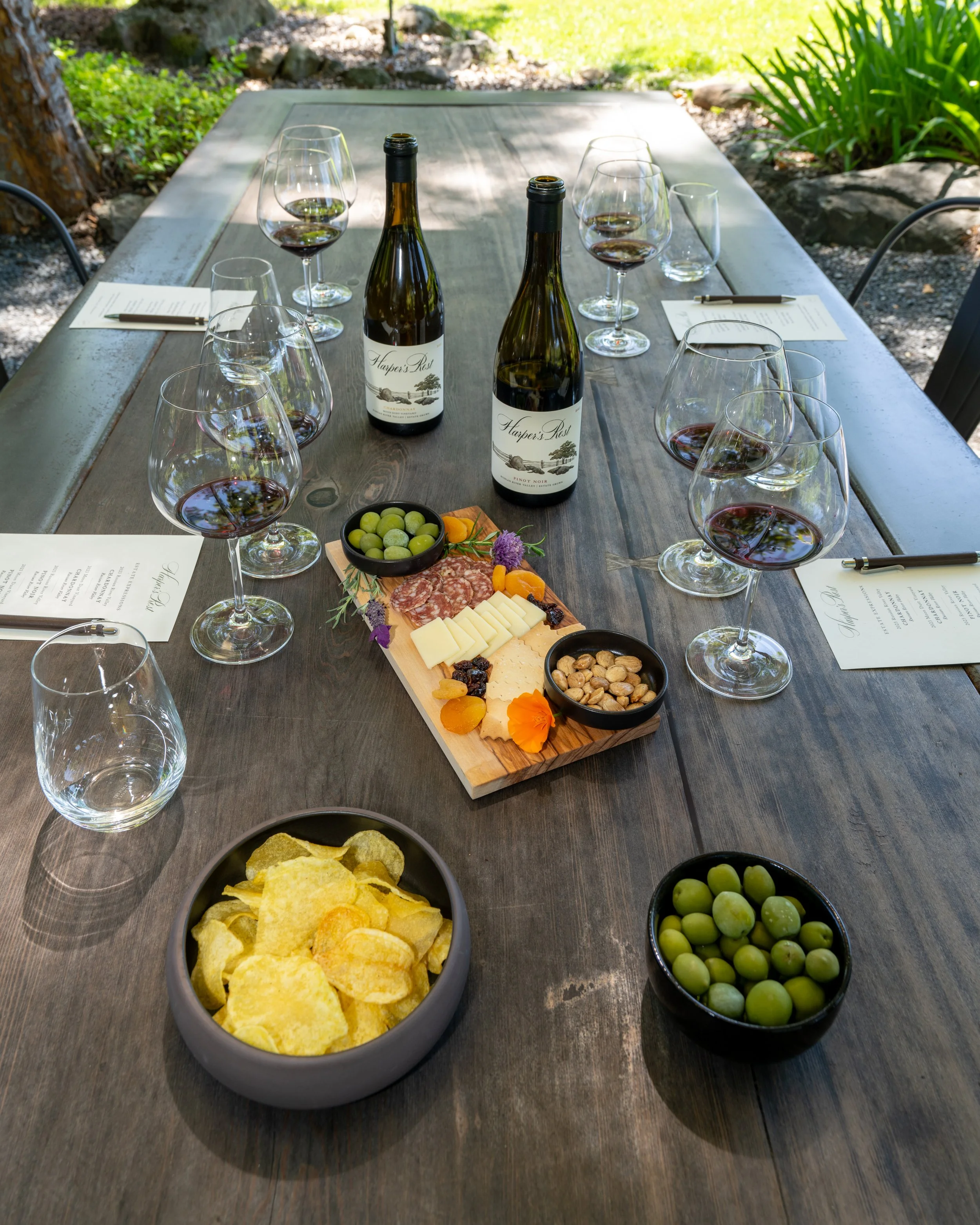 An outdoor wine tasting table set with Harper’s Rest Chardonnay and Pinot Noir, tasting glasses, notes, and a charcuterie board with cheese, salami, olives, nuts, dried fruit, and potato chips.