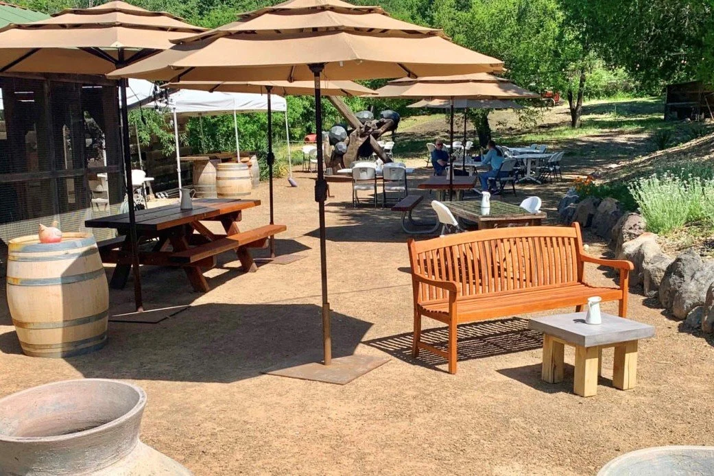 Outdoor patio area with umbrellas, wooden benches, tables, and people sitting and socializing surrounded by trees and rocks.