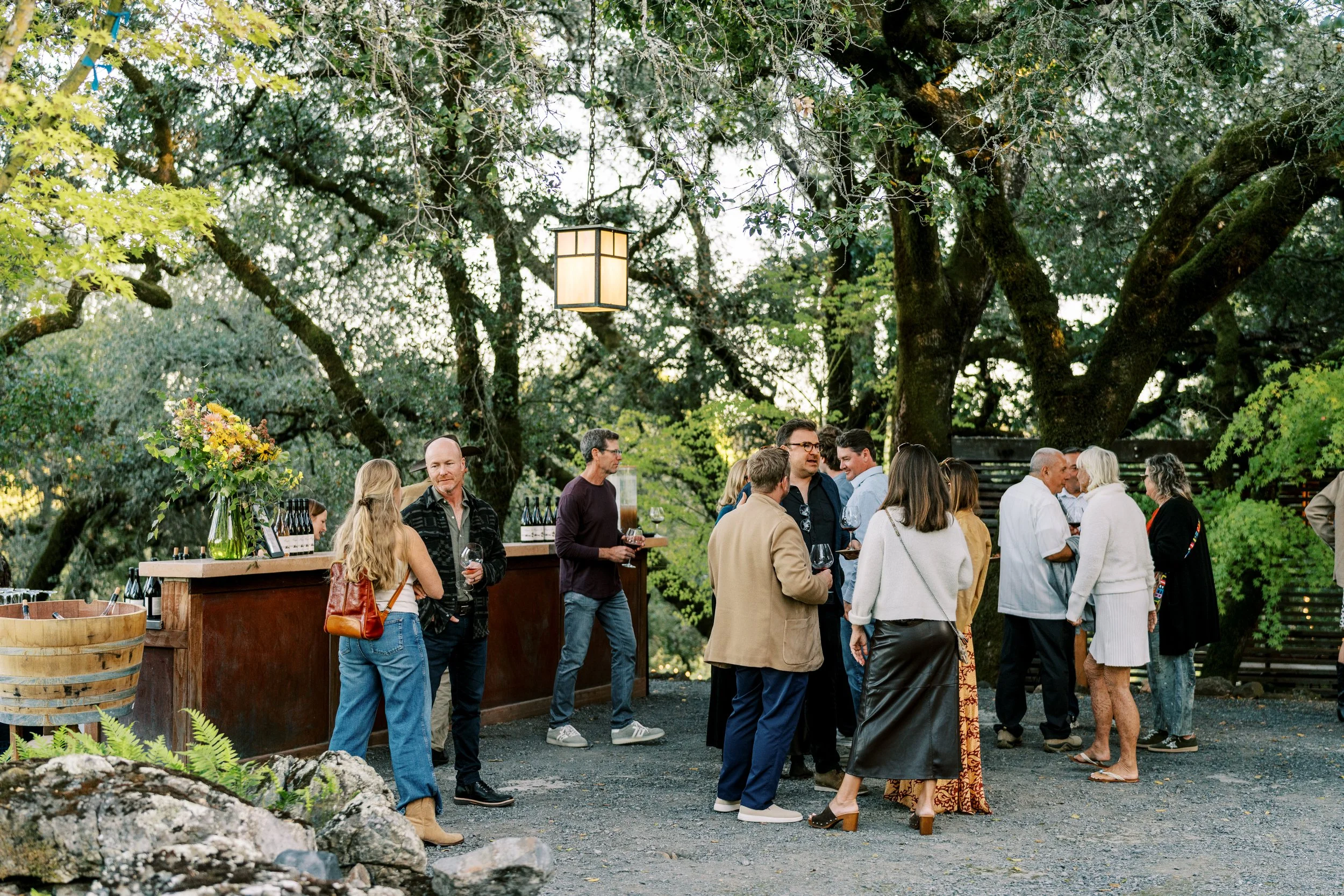 People socializing outdoors during daytime, with a bar area and large trees overhead.