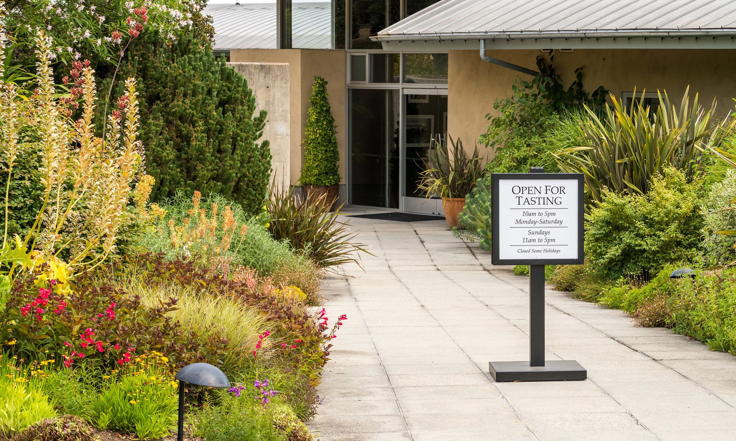 A walkway leading to a building with large glass entrance doors, surrounded by lush green plants and flowers. A sign in front indicates the place is open for tasting from 10 am to 5 pm Monday through Saturday, and 11 am to 5 pm on Sundays, with some 