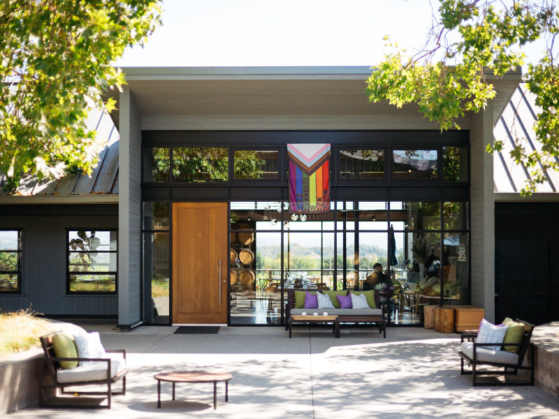 Modern building with large glass windows, outdoor seating area with chairs and cushions, trees, and a colorful banner inside.