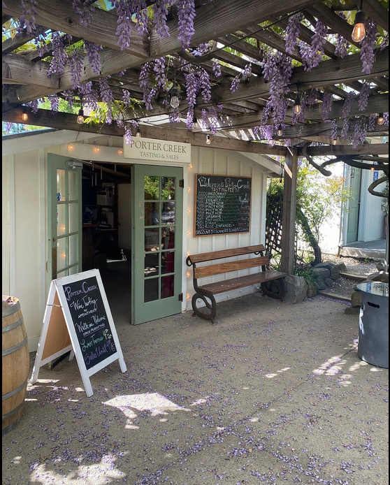 Exterior of a small tasting shop with a light green door, blackboard menus, and a wooden bench under a pergola decorated with purple wisteria flowers.