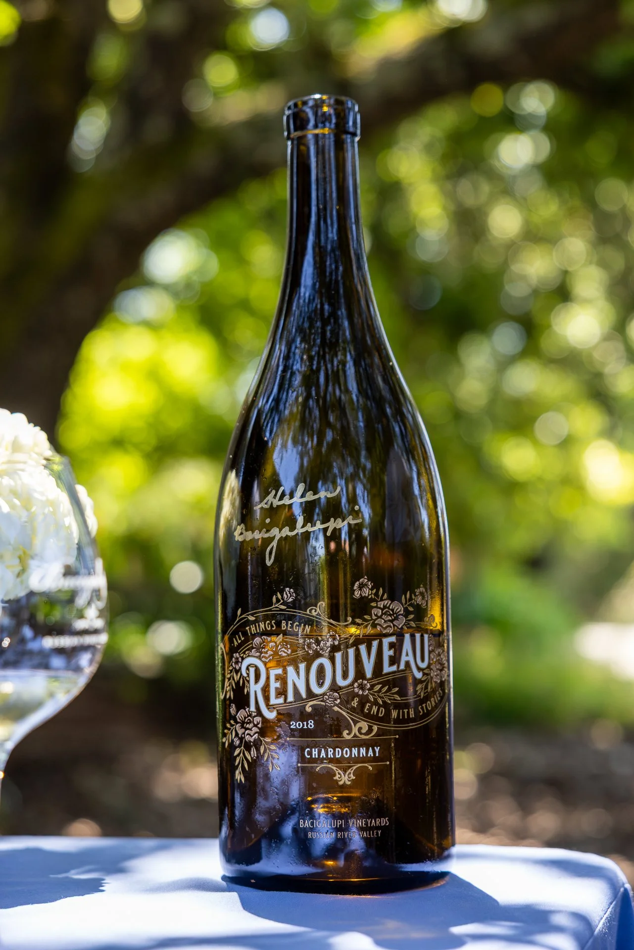 A bottle of Renouveau Chardonnay wine from Bacigalupi Vineyards, Russian River Valley, 2018, placed outdoors with greenery and sunlight in the background.