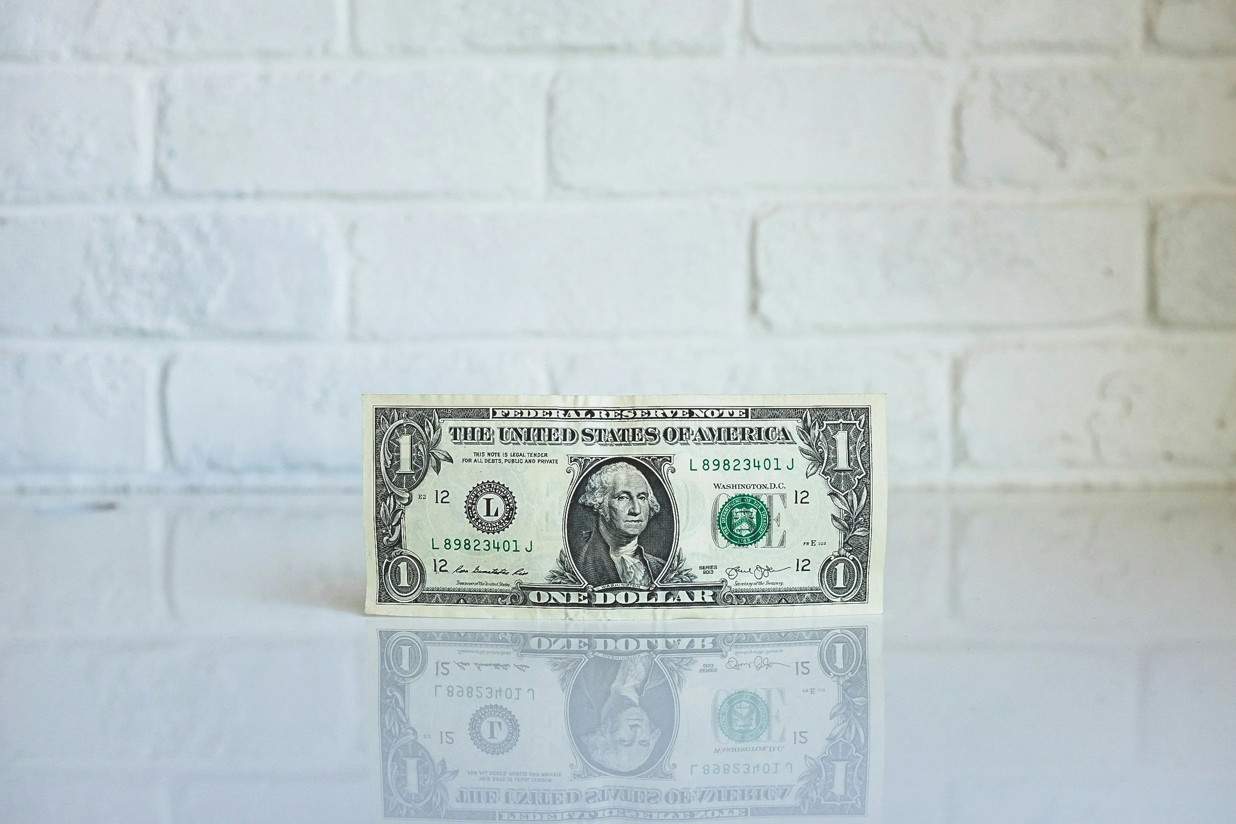 A one-dollar bill placed on a reflective surface with a white brick wall in the background.