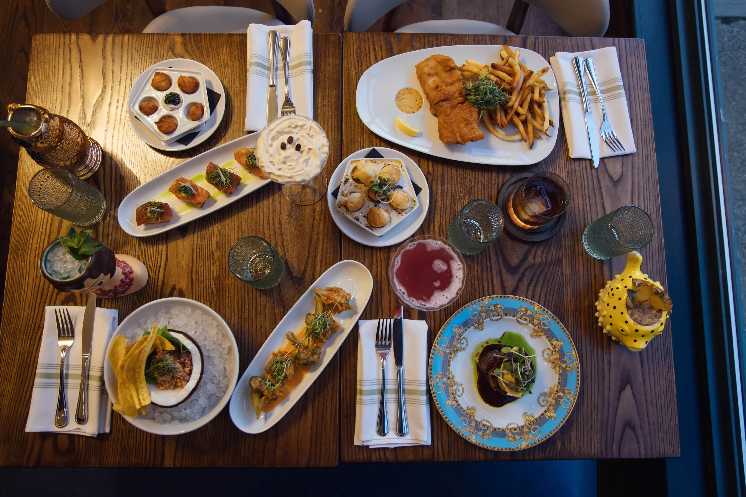 A top-down view of a wooden table set with various plates of food, drinks, and utensils, including fried fish and fries, sushi, appetizers, and desserts, with cloth napkins and glasses.