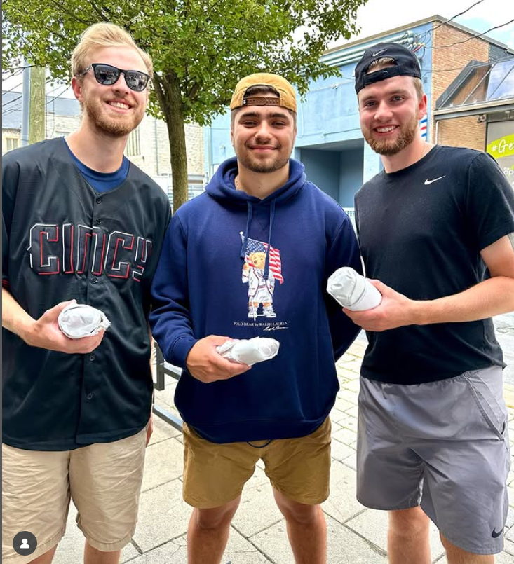 Three young men standing outdoors, each holding wrapped food items. The man on the left wears sunglasses and a black shirt with the word 'CUCH' on it. The man in the middle wears a navy hoodie with a graphic and a tan cap. The man on the right wears