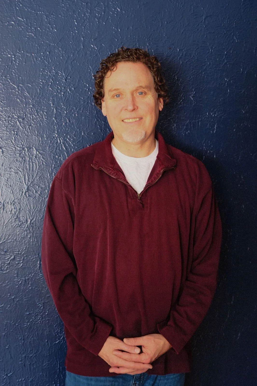 Original Medicine - Michael Frisby with curly brown hair, light skin, and a white goatee, wearing a maroon zip-up fleece jacket and a white shirt, standing in front of a textured dark blue wall.