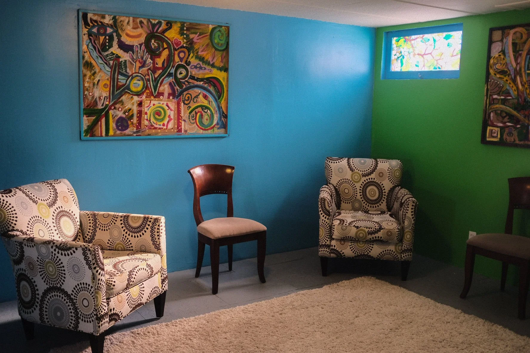 Original Medicine - A colorful living room with a blue and green wall, abstract paintings, a window with stained glass, patterned armchairs, a wooden chair, and a cream-colored rug.
