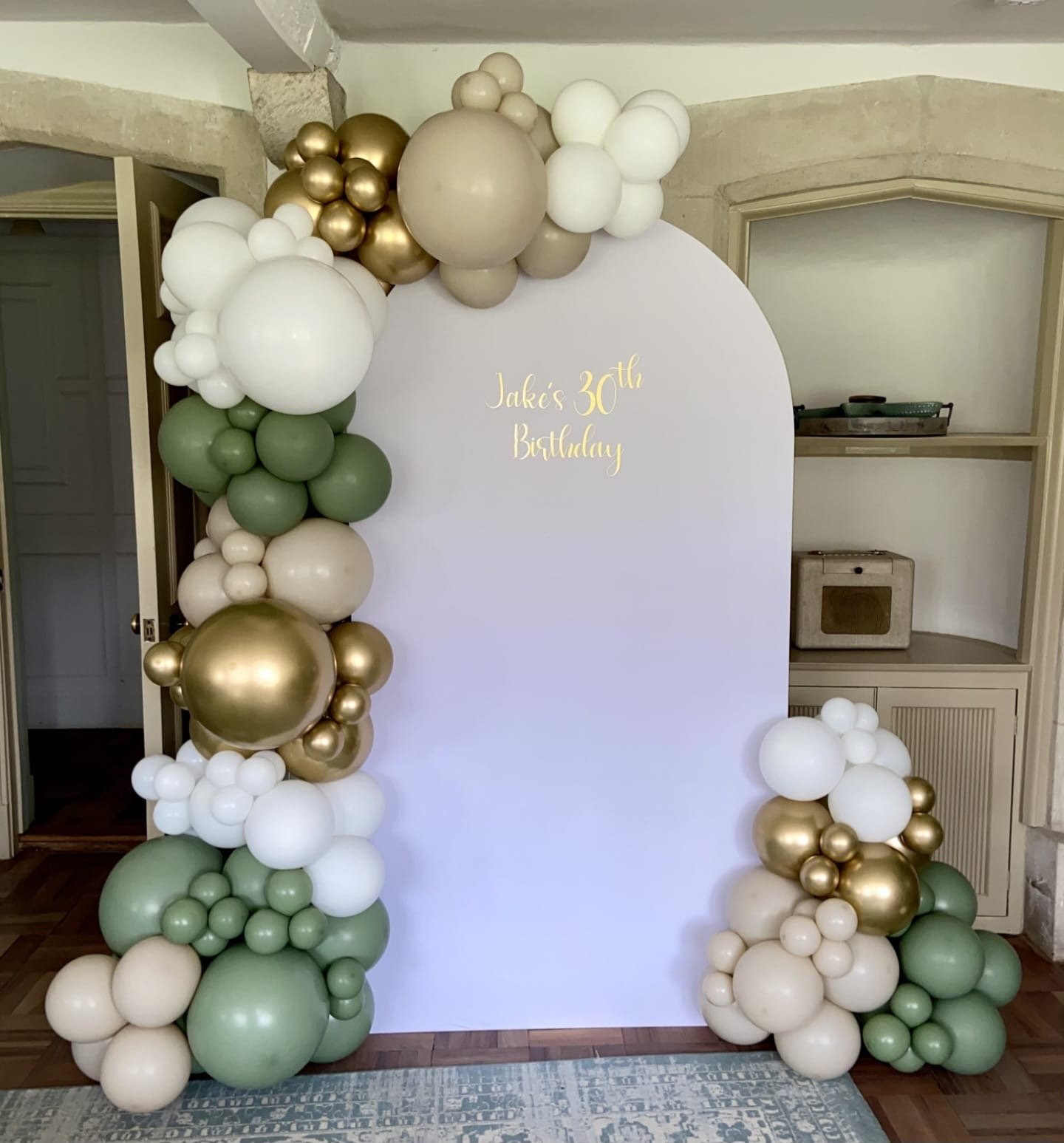 Balloon arch with white, green, gold, and beige balloons framing a white backdrop with gold lettering that reads "Jake's 30th Birthday".