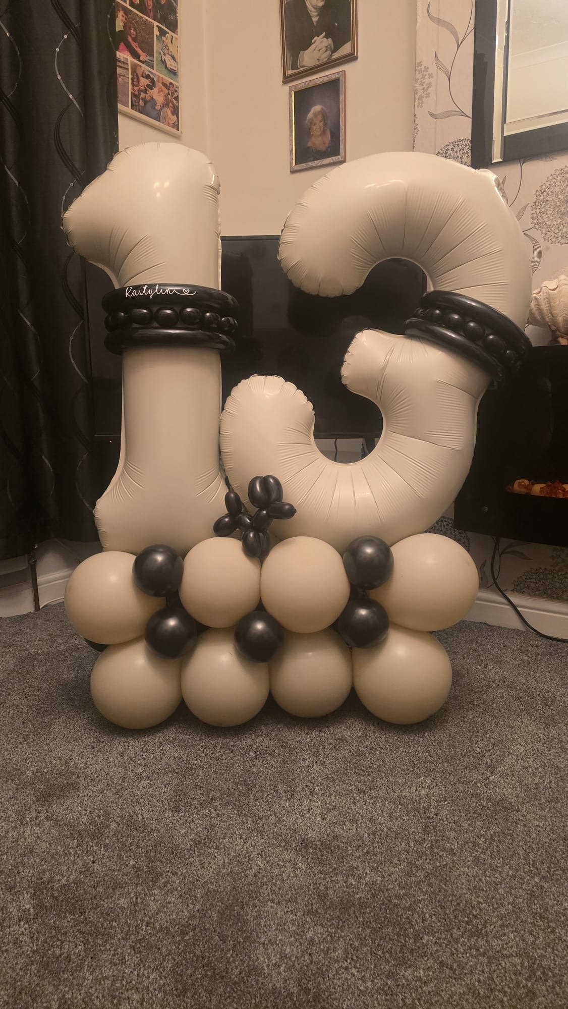 Large balloon arrangement decorated with black and white balloons forming the letters 'J'. The balloons are arranged base down on the carpeted floor, with decorative black balloons on the top and inside the letter. The background includes framed phot