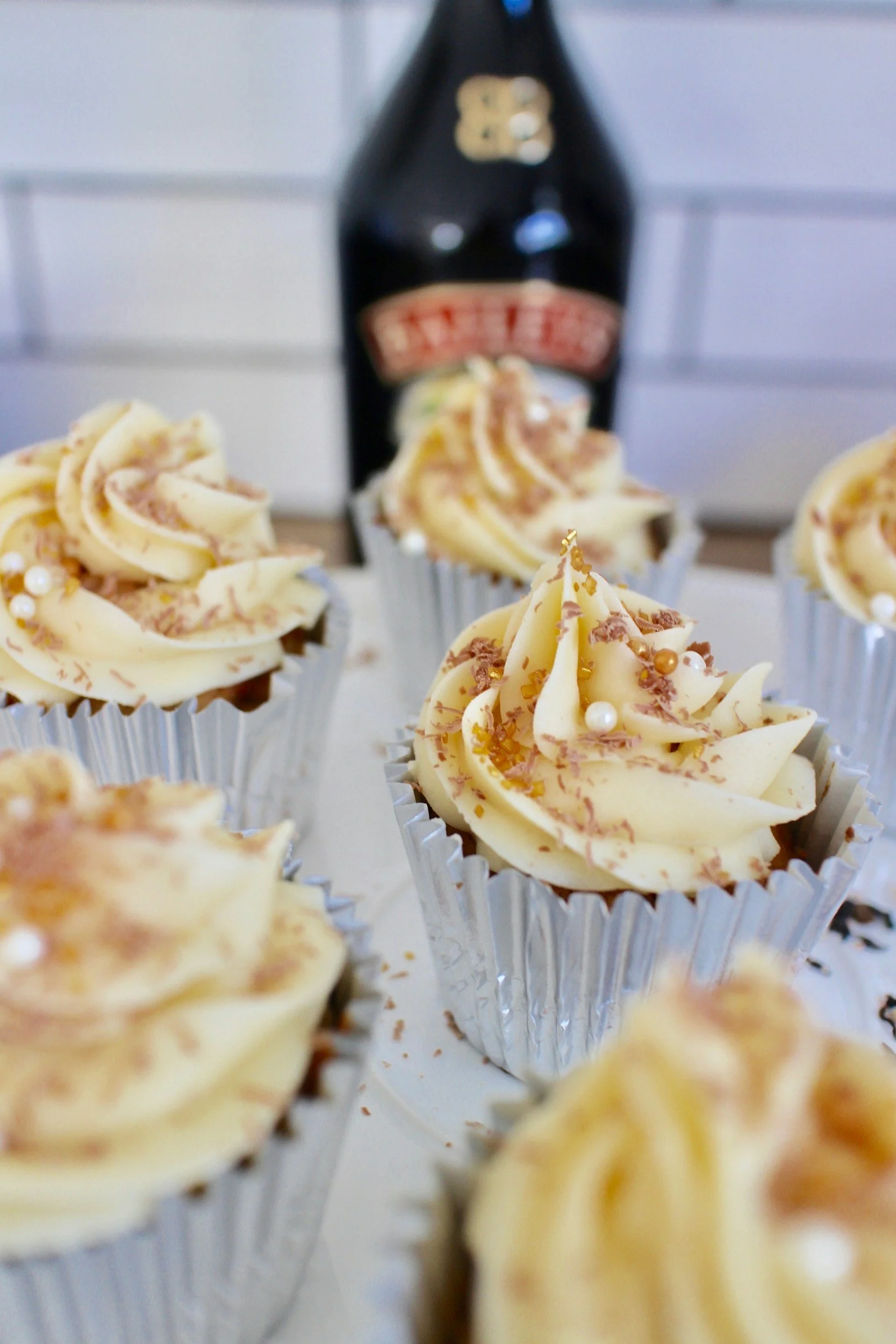 Cupcakes topped with white frosting, chocolate shavings, and pearl sprinkles, with a bottle of beer in the background.