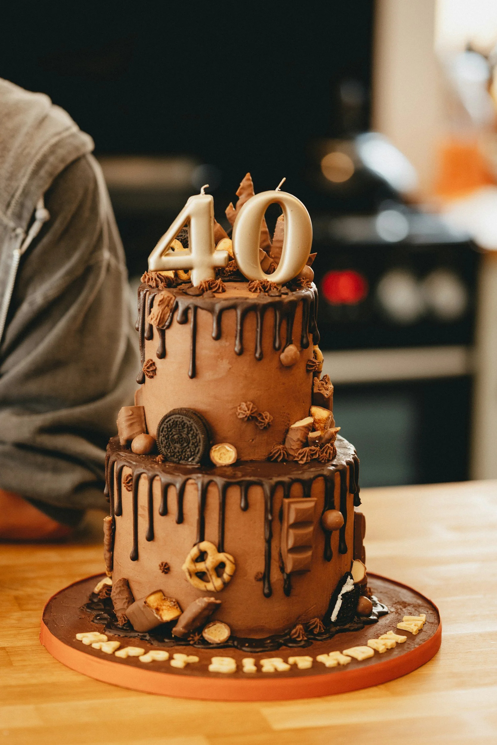 A two-tier chocolate birthday cake decorated with chocolates, cookies, and chocolate drips, with a gold '40' candle on top, on a wooden table.