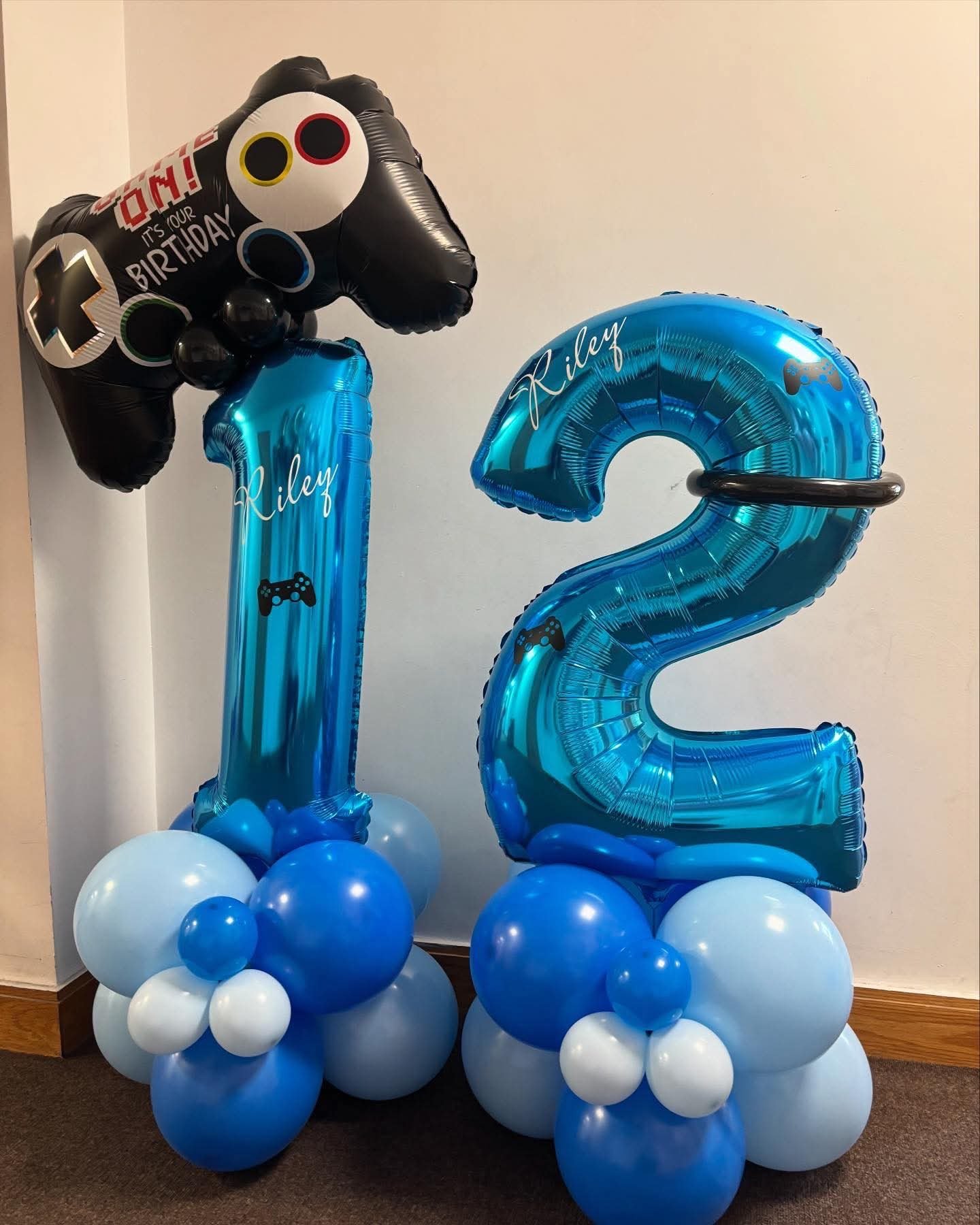 Blue number balloons with the name Riley, decorated with gaming controller graphics, topped by a black balloon shaped like a video game controller with a "It's Your Birthday!" message, arranged with additional blue, white, and light blue balloons at