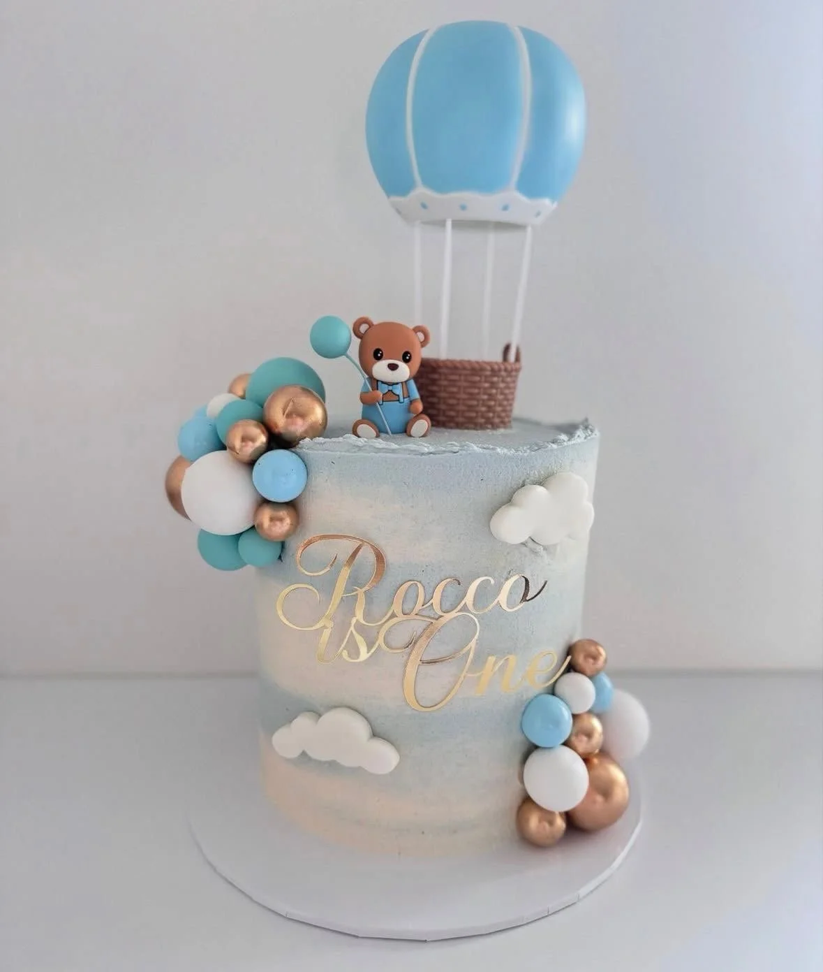 Decorative cake with clouds, balloons, teddy bear, hot air balloon topper, and a sign reading 'Rocco One' for a first birthday celebration.