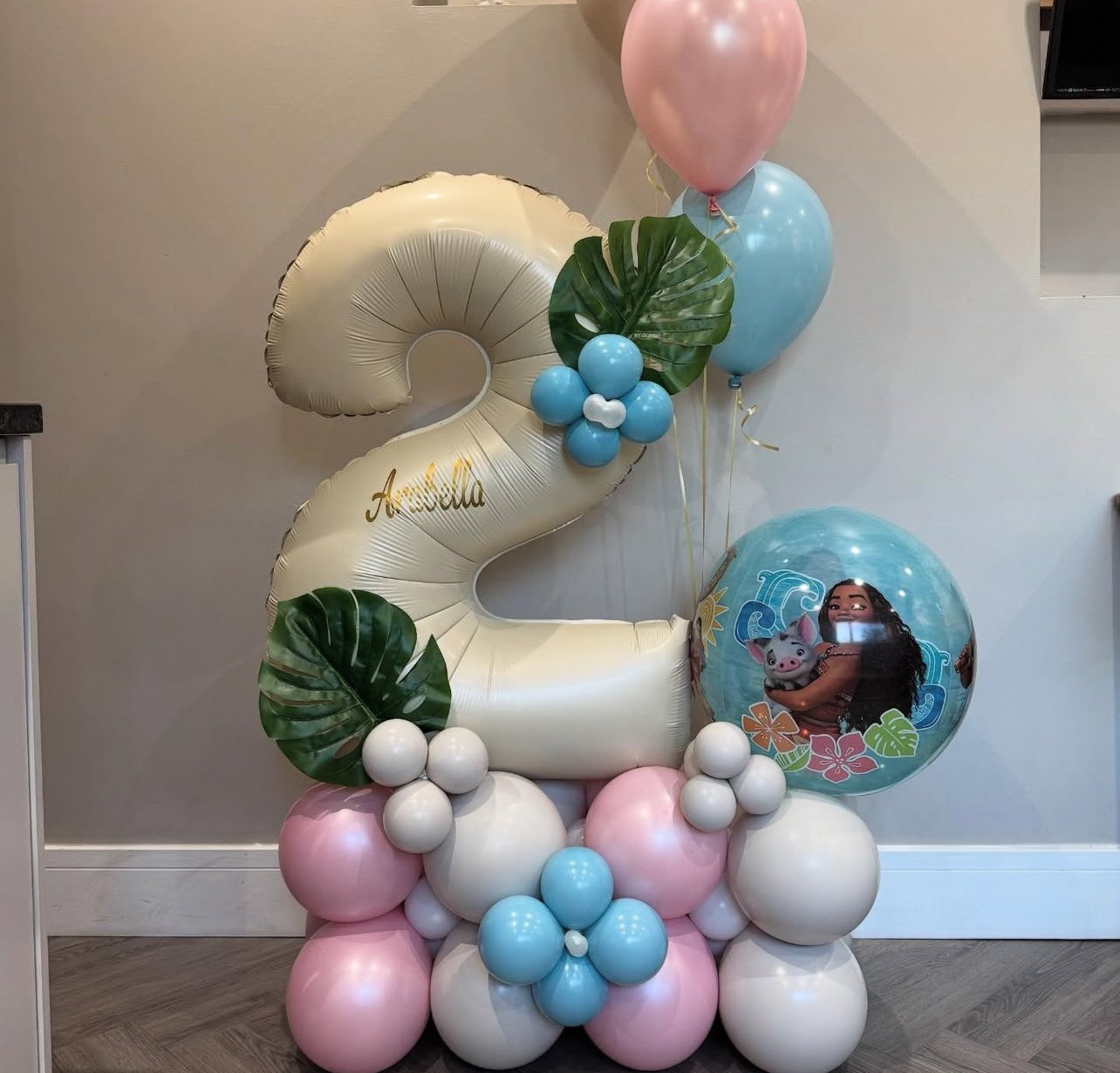A balloon arrangement celebrating a second birthday with a large cream number 2 balloon, pastel pink and blue balloons, green tropical leaves, a custom balloon with a character and flowers, and the name Amabella written in gold.