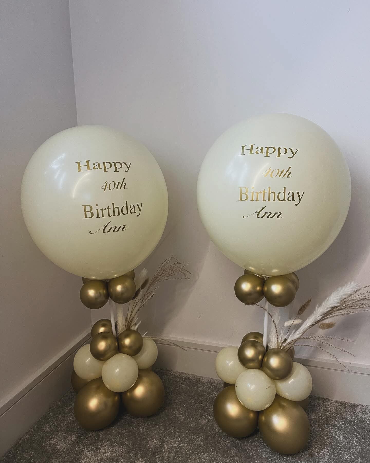 Two large cream-colored balloons with gold and black lettering celebrating Ann's 40th birthday, anchored by clusters of gold and cream balloons at the base, with decorative dried grasses decorating the sides.
