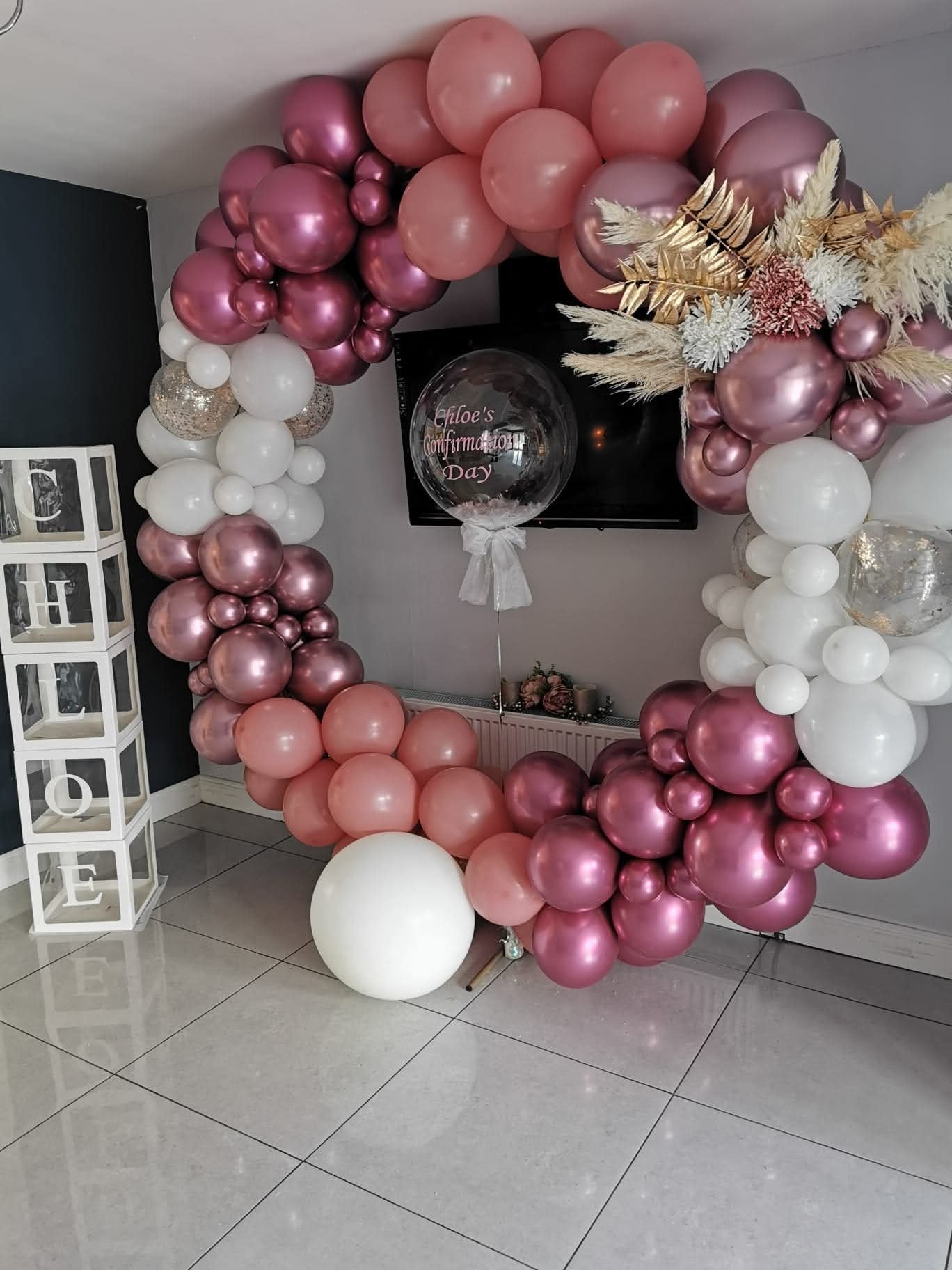 A circular balloon arch decorated with pink, white, and purple balloons, with a clear balloon in the center that reads 'Chloe's Confirmation Day' in pink. The arch is set up in front of a wall-mounted TV, with a white stand with the word 'LOVE' on th