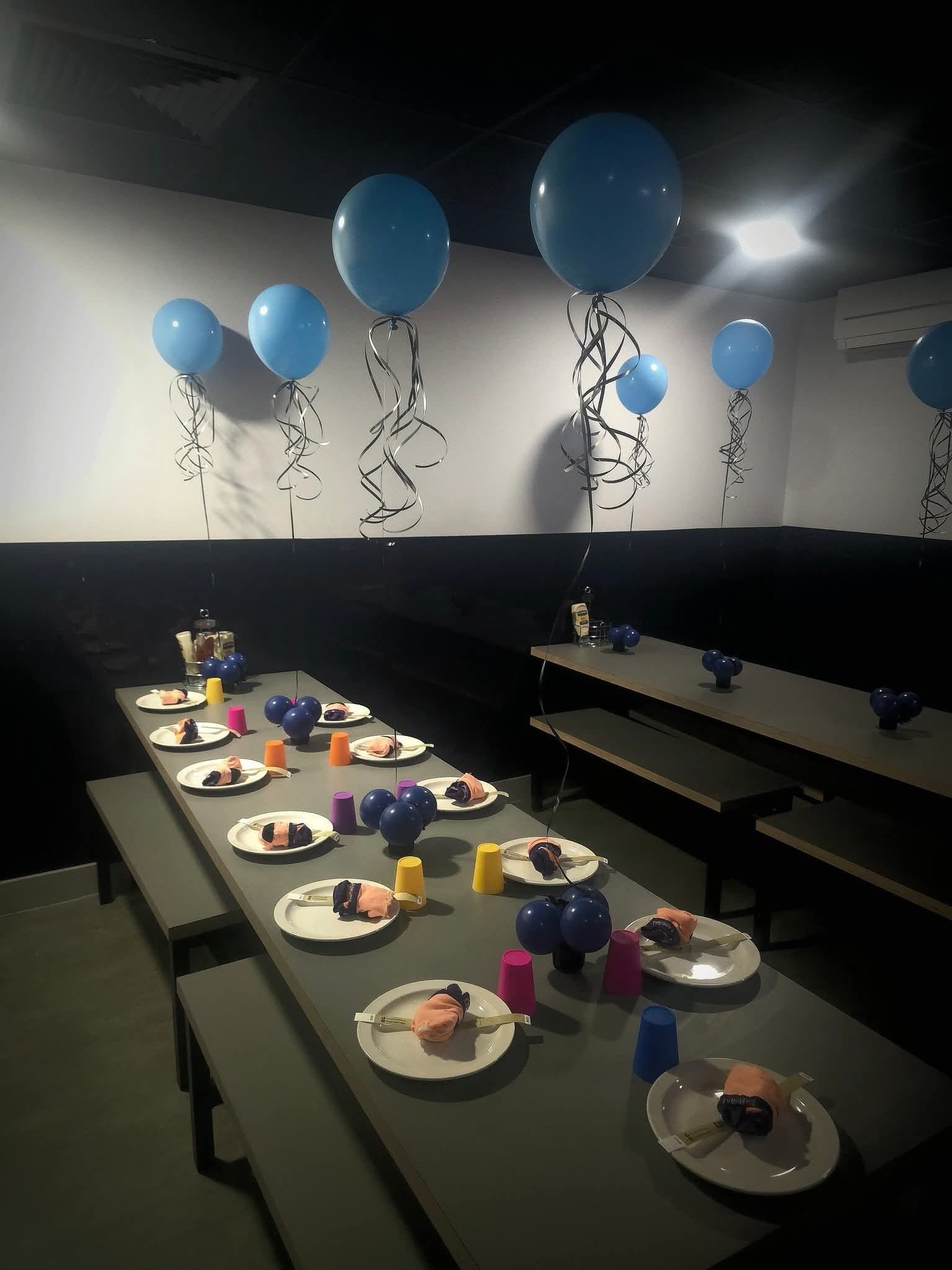 Party table setup with blue balloons, colorful cups, and plates with wrapped candies, in a dimly lit room.