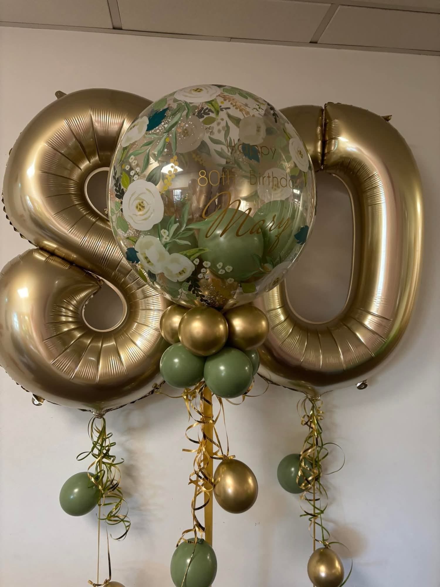Gold and green balloons arranged for a birthday celebration, including two large gold number balloons '8' and '0', and a transparent balloon with floral design and the message 'Happy 80th Birthday Mary.'