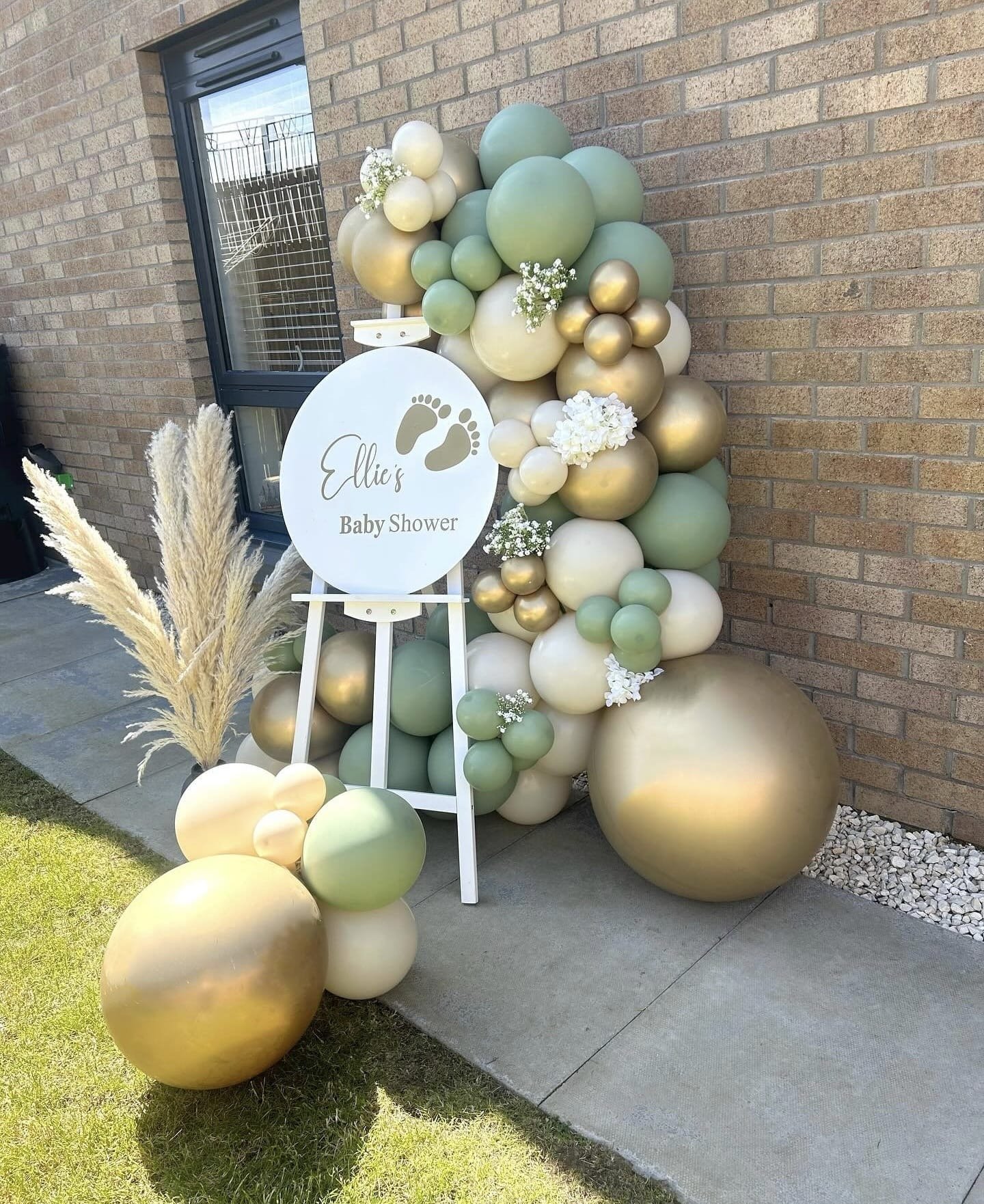 Decorative balloon arrangement for a baby's shower with a white sign reading 'Ellie's Baby Shower', featuring tan, white, green, and gold balloons along a brick wall, with pampas grass and small white flowers.