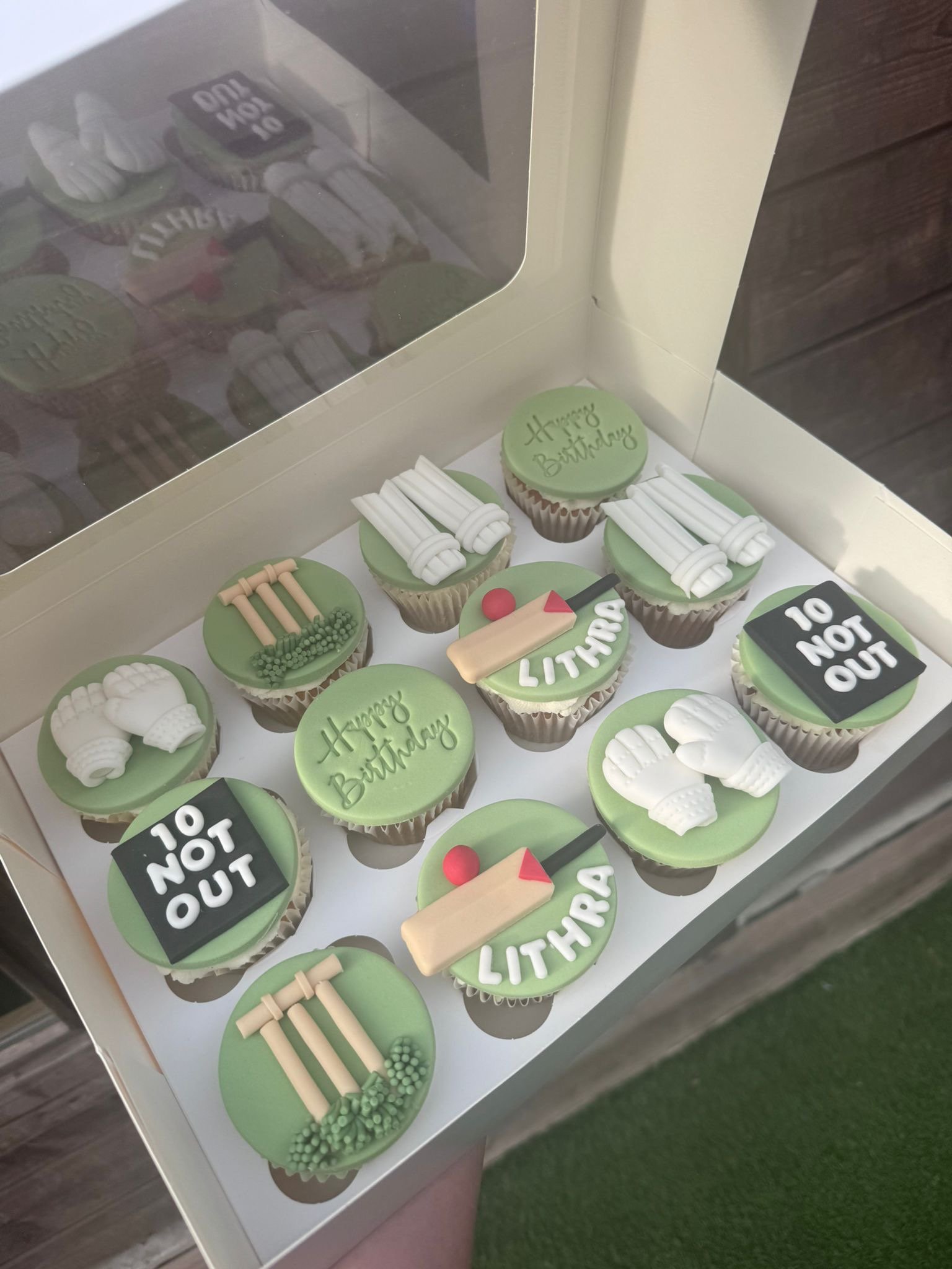 A box of cupcakes with golf-themed decorations including golf clubs, golf balls, and the words 'Happy Birthday' and 'LITHRA.' Some cupcakes have '10 NOT OUT' written on them.