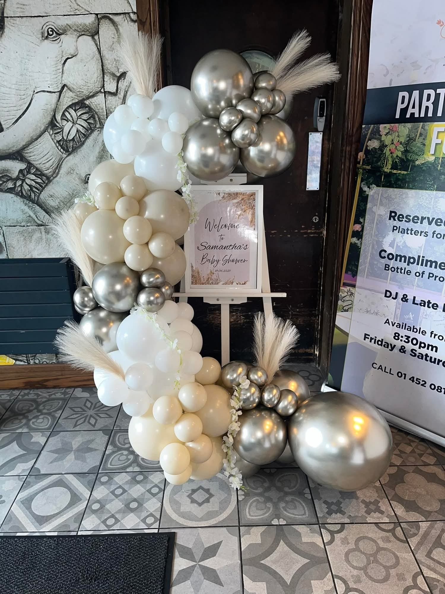 Balloon arch with white, cream, and metallic silver balloons, decorated with pampas grass and white flowers, at a baby shower event.