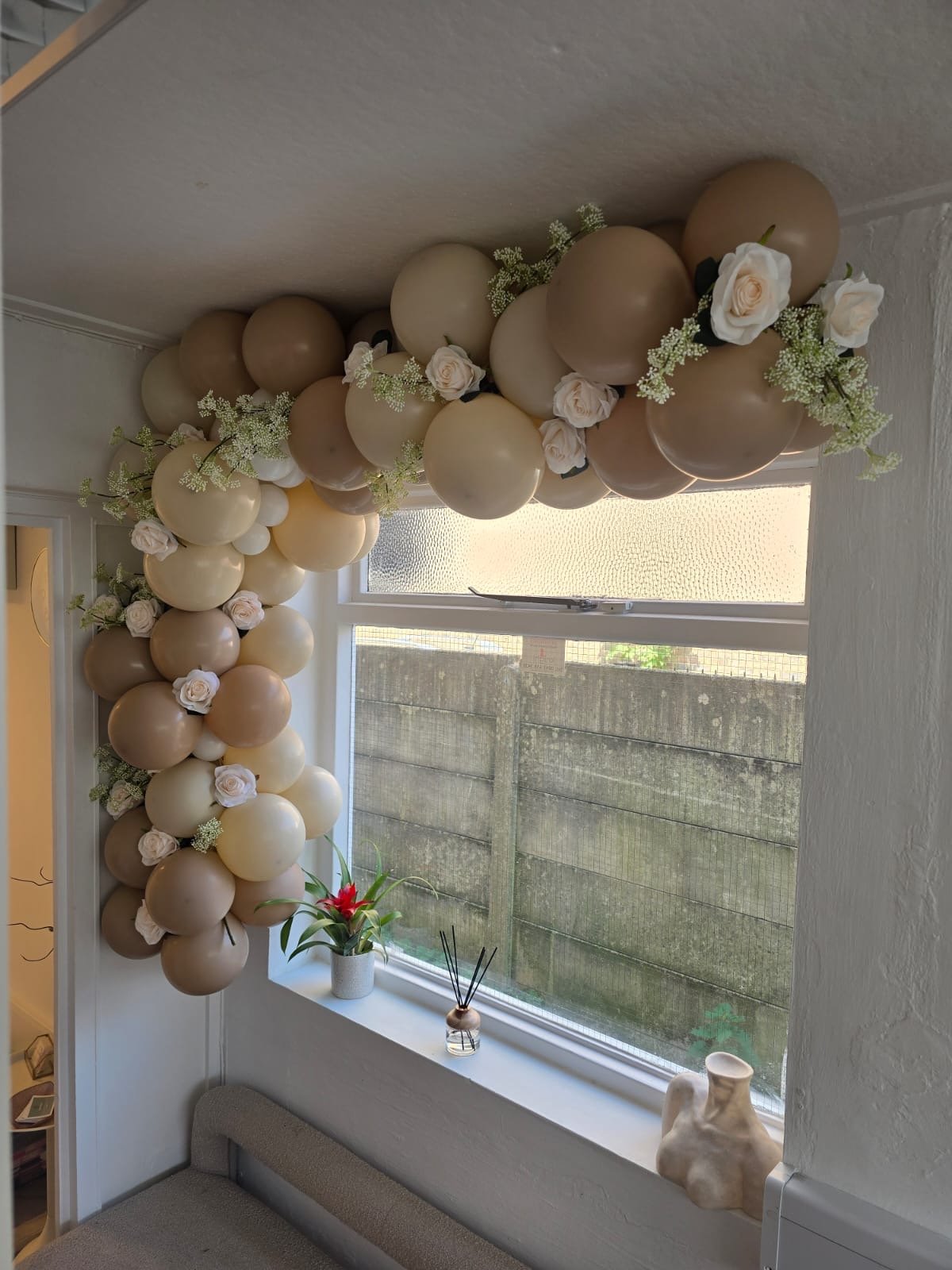 Balloon garland with white and beige balloons, white roses, and green foliage hanging above a window with a windowsill decorated with a potted plant, a reed diffuser, and a vase, in a cozy room.