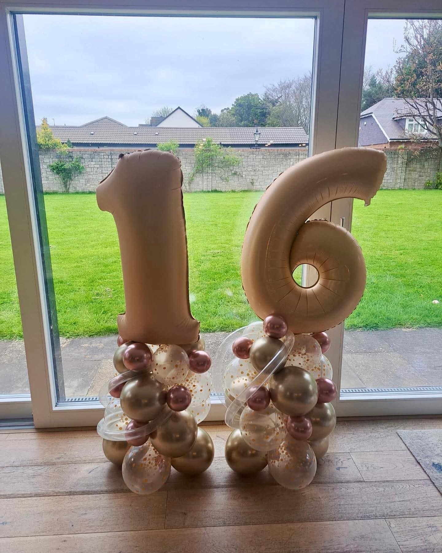 Large gold number balloons '1' and '6' with smaller balloons at their base, standing in front of a glass door, with a backyard and cloudy sky visible outside.
