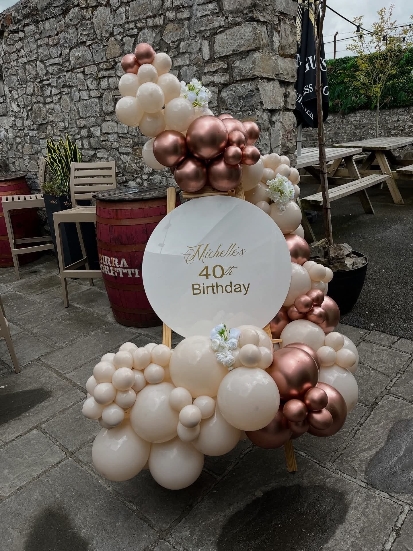 Decorative balloon arrangement with plaques celebrating Michelle's 40th birthday, set outdoors against a stone wall with chairs, tables, and potted plants surrounding it.