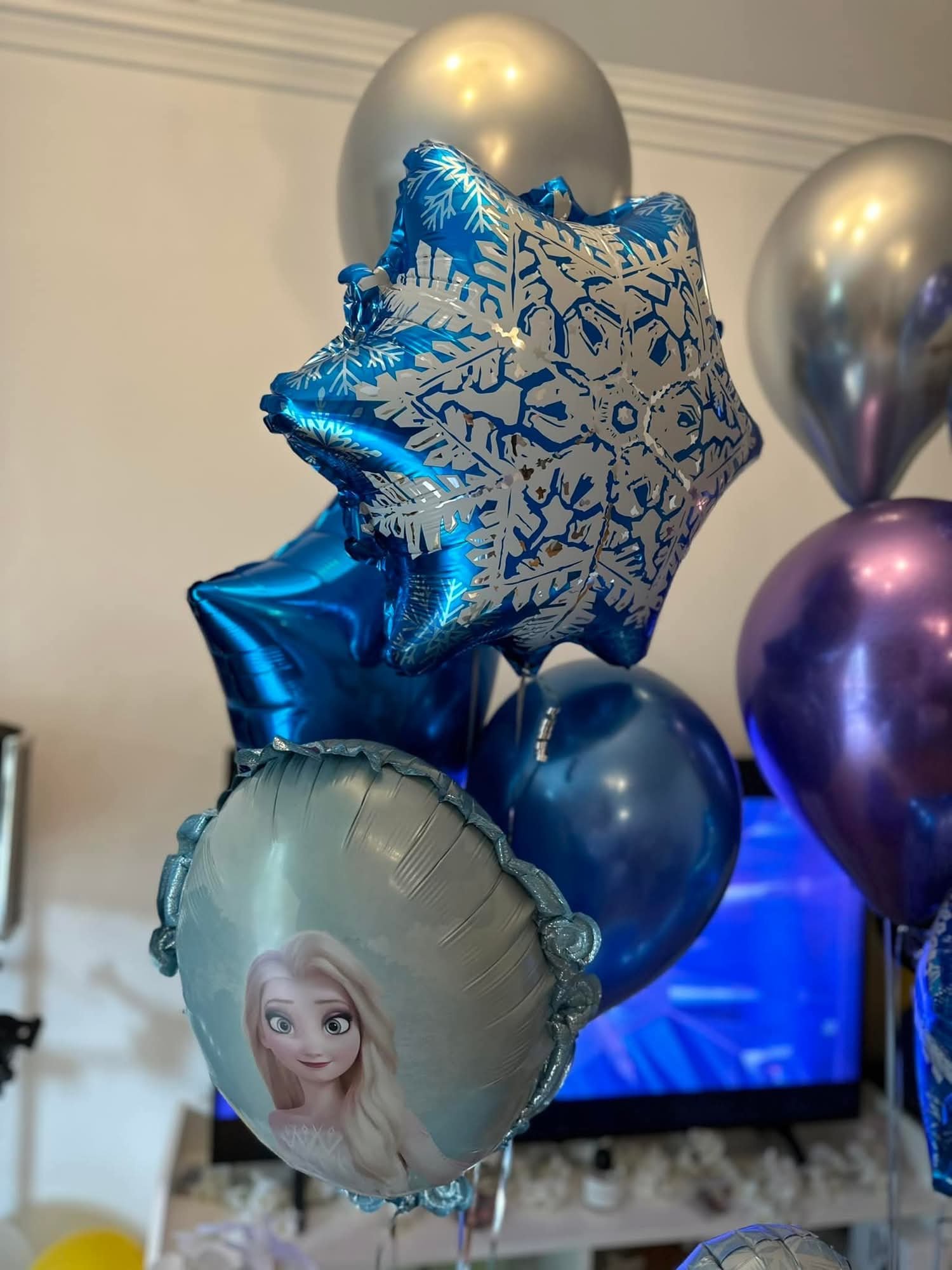 A cluster of balloons, including a large star-shaped blue balloon with snowflake designs, a round silver balloon with a character from Frozen, and other metallic balloons in purple, silver, and blue, set indoors.