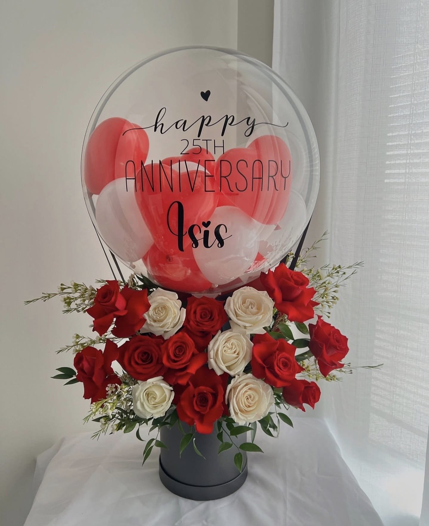 Flower arrangement with red and white roses in a black vase, topped with a transparent balloon reading 'happy 25th anniversary Isis' with red and white balloons inside.