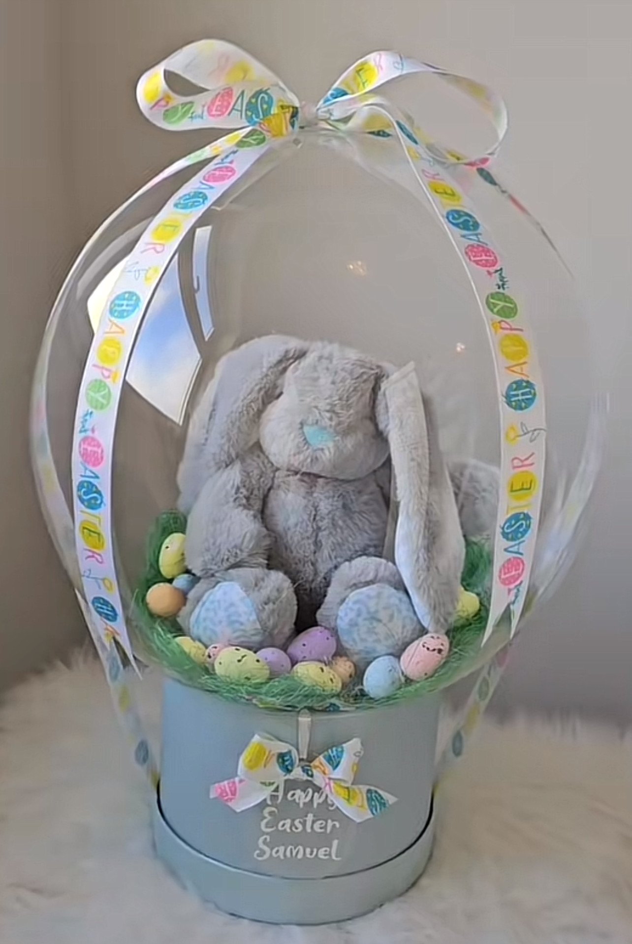 Easter basket with a plush gray bunny, pastel colored eggs, and decorative ribbon reading 'Happy Easter'.