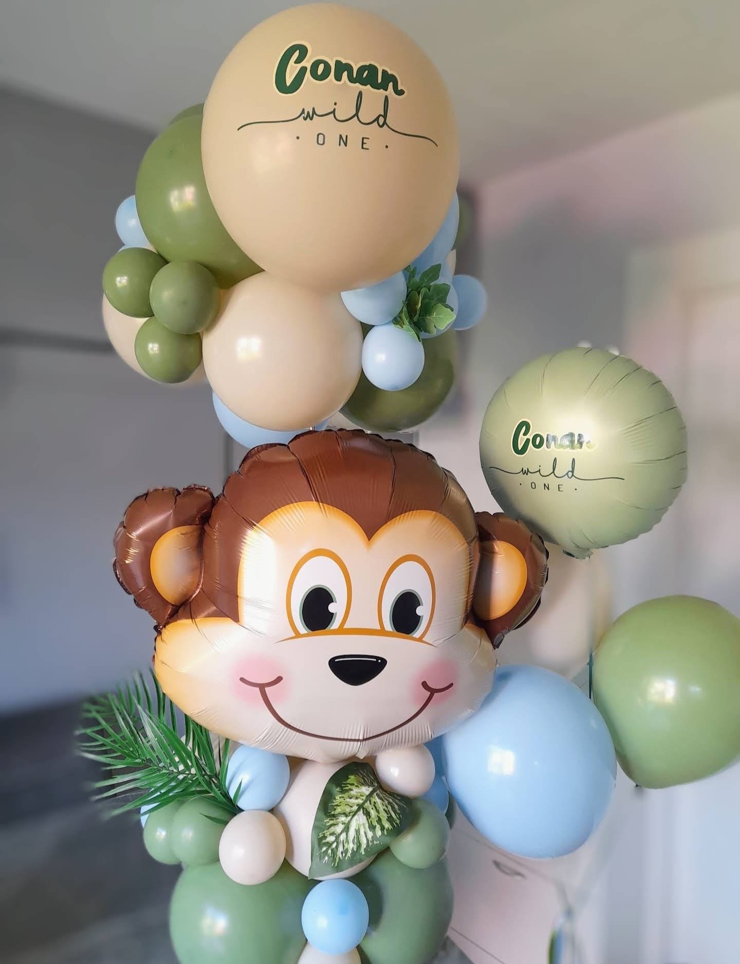 Balloon arrangement with a large monkey face balloon, green, white, and blue round balloons, and two beige balls with the text "Conan wild ONE."